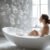 A woman with her hair in a bun relaxes in a bathtub filled with bubbles. The modern bathroom design is softly lit, and there are numerous bubbles floating in the air. She gazes towards a sunlit window in a serene and peaceful setting, reminiscent of an elegant London retreat.